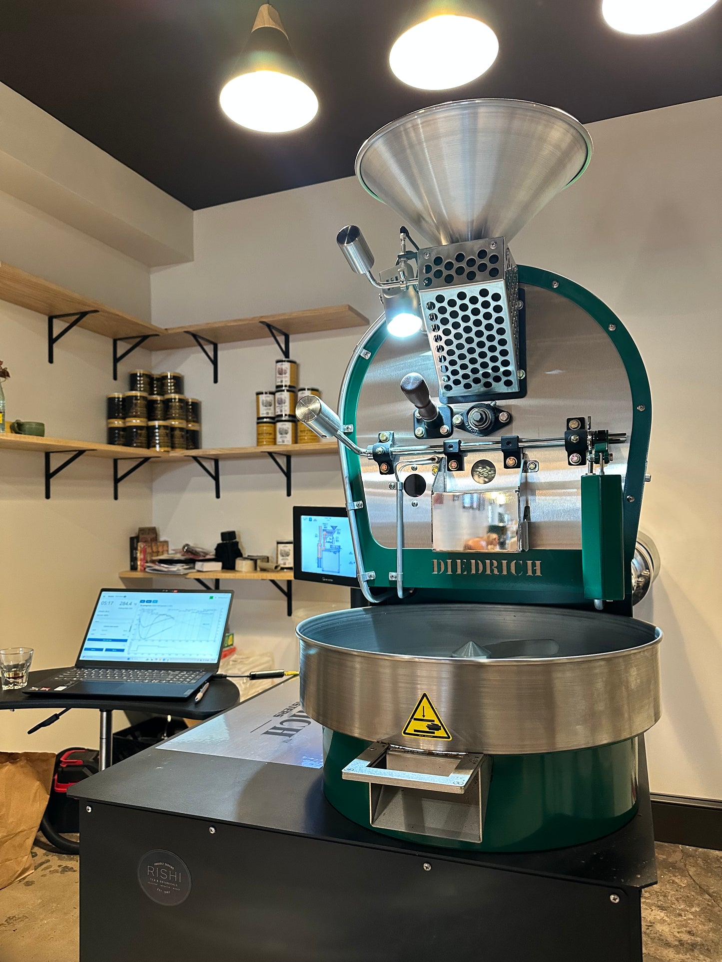 Coffee roaster in a workshop setting with shelves and computers in the background.
